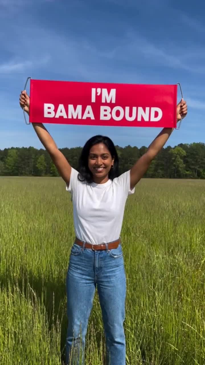 A woman stands in a sunny field, smiling, with a red I'M BAMA BOUND banner held flat overhead. The banner has no school logo. Not made by this studio.